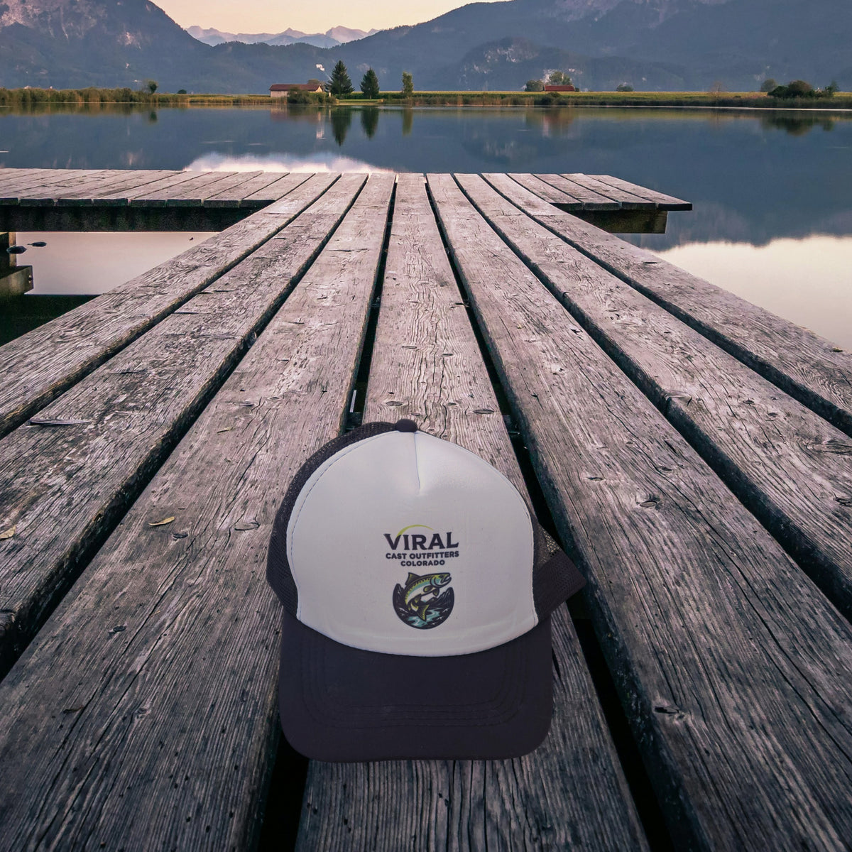 Viral Cast Outfitters black and white trucker hat on a wooden lake dock at dusk, mountains in the background