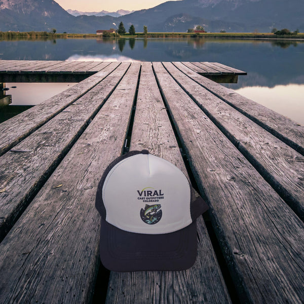 Viral Cast Outfitters black and white trucker hat on a wooden lake dock at dusk, mountains in the background