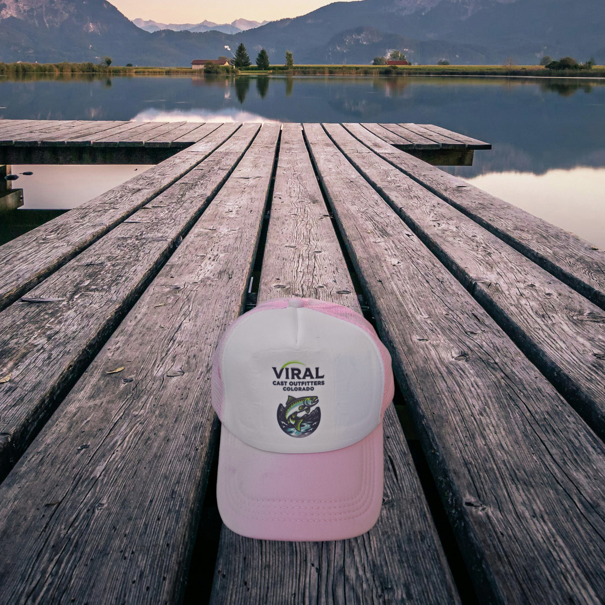 Viral Cast Outfitters pink and white trucker hat on a wooden lake dock at dusk, mountains in the background
