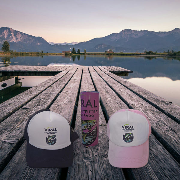 Viral Cast Outfitters black and white trucker hat, pink tumbler and pink and white trucker hat on a wooden lake dock at dusk